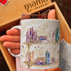 Pode incluir: Uma caneca de cer&acirc;mica branca com uma ilustra&ccedil;&atilde;o de contornos dourados de duas prateleiras cheias de livros coloridos em tons de roxo, azul e rosa. O design inclui delicadas vinhas e caules florais dourados ao redor dos livros.