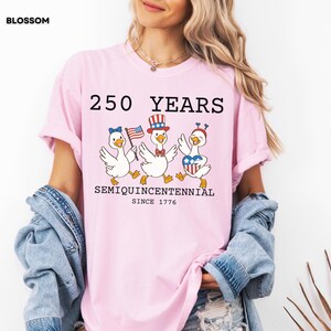 May include: A light pink t-shirt with the text "250 YEARS SEMIQUICENTENNIAL SINCE 1776" and illustrations of three cartoon geese in patriotic attire. The geese are wearing a bow, a hat, and a heart-shaped design.