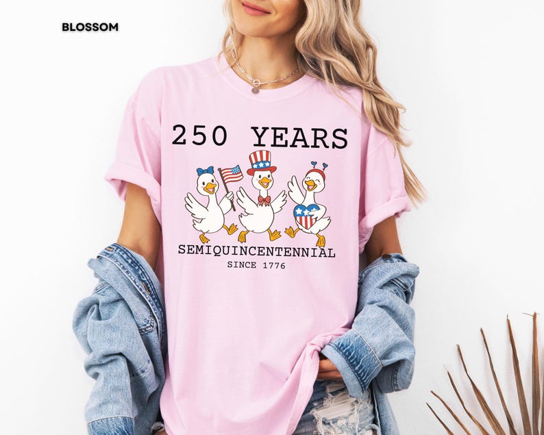 May include: A light pink t-shirt with the text "250 YEARS SEMIQUICENTENNIAL SINCE 1776" and illustrations of three cartoon geese in patriotic attire. The geese are wearing a bow, a hat, and a heart-shaped design.