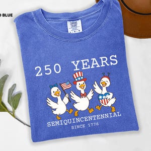 May include: Flo blue t-shirt with the text "250 YEARS SEMIQUINCENTENNIAL SINCE 1776". The shirt features a graphic of three cartoon ducks wearing patriotic accessories, including a hat, bow, and heart-shaped emblem.