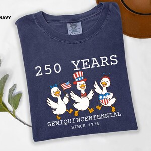 May include: Navy blue t-shirt with a graphic celebrating 250 years since 1776. The design features three cartoon ducks wearing patriotic accessories and holding flags. The text reads "250 YEARS SEMIQUINCENTENNIAL SINCE 1776."