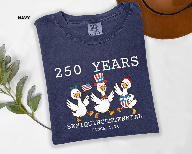 May include: Navy blue t-shirt with a graphic celebrating 250 years since 1776. The design features three cartoon ducks wearing patriotic accessories and holding flags. The text reads "250 YEARS SEMIQUINCENTENNIAL SINCE 1776."