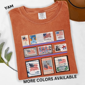 May include: A burnt orange t-shirt featuring a collection of vintage-style postage stamps with American flag designs. The shirt includes the text "YAM" at the top left and "MORE COLORS AVAILABLE" at the bottom. The shirt is a Comfort Colors brand.