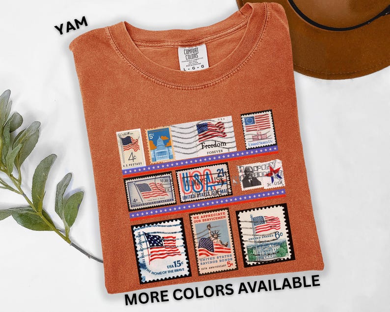 May include: A burnt orange t-shirt featuring a collection of vintage-style postage stamps with American flag designs. The shirt includes the text "YAM" at the top left and "MORE COLORS AVAILABLE" at the bottom. The shirt is a Comfort Colors brand.