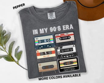 May include: Pepper-colored t-shirt with the text "IN MY 90'S ERA" and a graphic of several cassette tapes. The shirt also has the text "MORE COLORS AVAILABLE". The shirt is made of a soft, comfortable material.