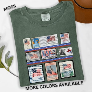 May include: Moss green t-shirt featuring a collection of vintage-style postage stamps with American flag designs. The stamps are arranged in rows and include the text "Freedom Forever" and "USA". The shirt also has the text "MORE COLORS AVAILABLE".