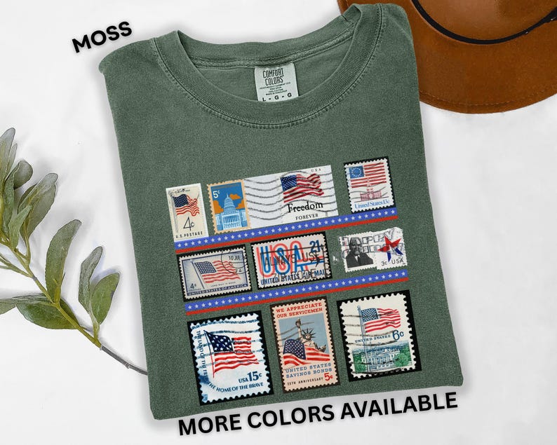 May include: Moss green t-shirt featuring a collection of vintage-style postage stamps with American flag designs. The stamps are arranged in rows and include the text "Freedom Forever" and "USA". The shirt also has the text "MORE COLORS AVAILABLE".