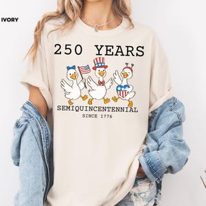 May include: Ivory-colored t-shirt featuring a graphic of three cartoon geese celebrating with patriotic accessories. The text reads "250 YEARS SEMIQUINCENTENNIAL SINCE 1776". The geese are wearing hats and holding flags, celebrating the anniversary.