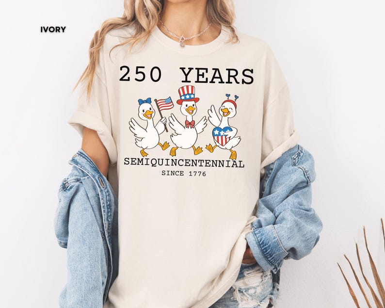 May include: Ivory-colored t-shirt featuring a graphic of three cartoon geese celebrating with patriotic accessories. The text reads "250 YEARS SEMIQUINCENTENNIAL SINCE 1776". The geese are wearing hats and holding flags, celebrating the anniversary.