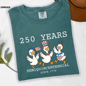 May include: Blue spruce Comfort Colors t-shirt with a graphic celebrating 250 years. The design features three cartoon ducks wearing patriotic hats and holding flags. Text includes "250 YEARS" and "SEMIQUINCENTENNIAL SINCE 1776".