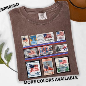 May include: Espresso-colored t-shirt featuring a collection of vintage-style postage stamps with American flag designs. The stamps are arranged in rows with red and blue accents. The text "MORE COLORS AVAILABLE" is at the bottom.