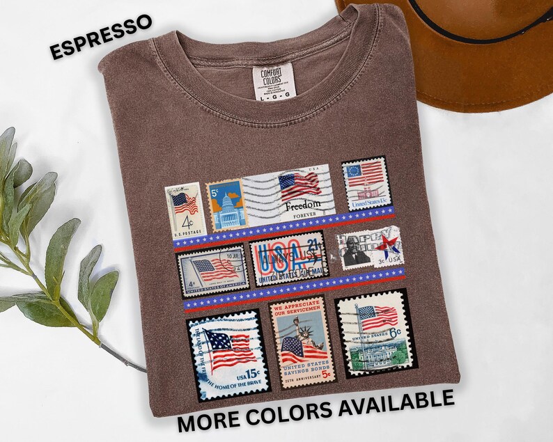 May include: Espresso-colored t-shirt featuring a collection of vintage-style postage stamps with American flag designs. The stamps are arranged in rows with red and blue accents. The text "MORE COLORS AVAILABLE" is at the bottom.
