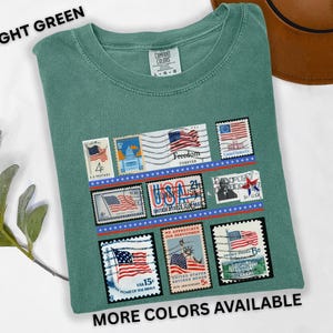 May include: Light green t-shirt featuring a collection of vintage-style postage stamps with American flag designs. The shirt includes the text "LIGHT GREEN" and "MORE COLORS AVAILABLE". The stamps are arranged in rows with red and blue borders.