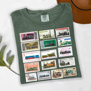 May include: Green Comfort Colors t-shirt with a collection of colorful postage stamps featuring various train designs. The stamps are arranged in a grid pattern on the front of the shirt. The shirt has a crew neck and short sleeves.