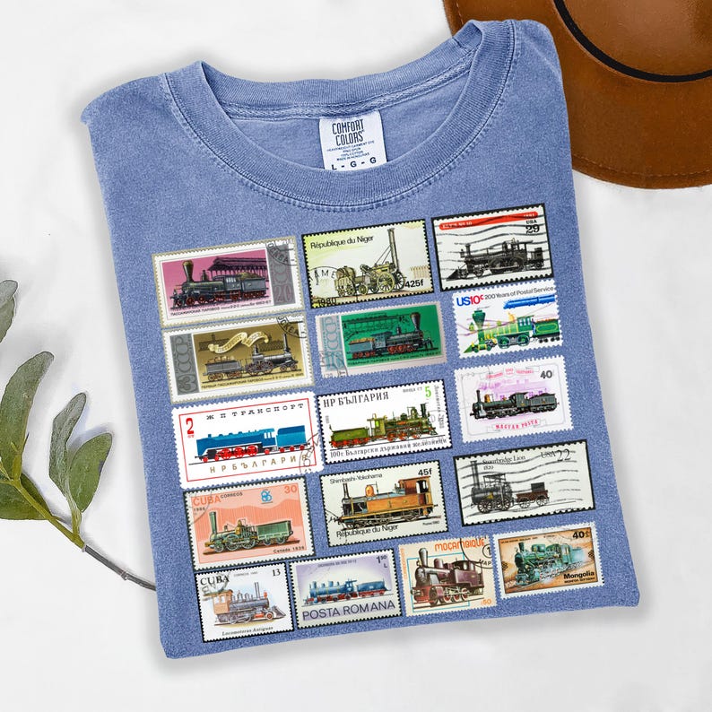 May include: A light blue Comfort Colors t-shirt featuring a collection of vintage train postage stamps. The stamps showcase various train designs in colors like green, blue, and brown. The shirt has a classic crew neck and short sleeves.
