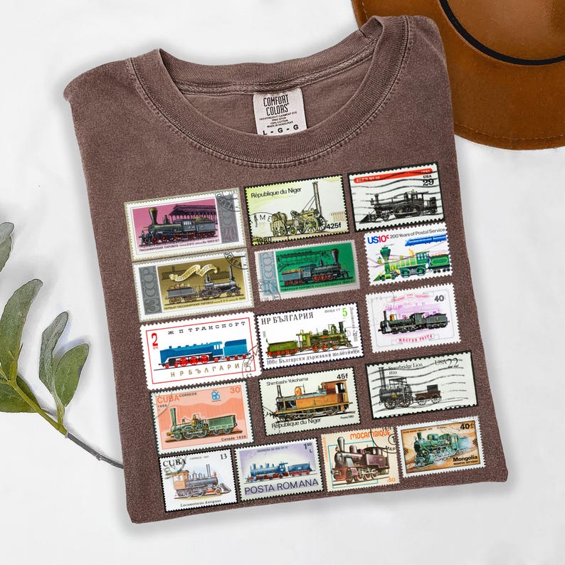 May include: Brown Comfort Colors t-shirt featuring a collection of vintage postage stamps with train illustrations. The stamps are in various colors and sizes, with text in multiple languages. The shirt has a crew neck and a tag that reads "Comfort Colors".