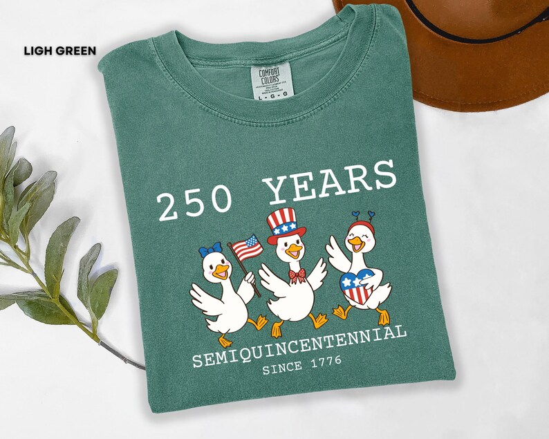 May include: Light green t-shirt with the text "250 YEARS SEMIQUINCENTENNIAL SINCE 1776". The shirt features a graphic of three cartoon ducks wearing patriotic hats and holding flags. A brown hat is visible in the background.