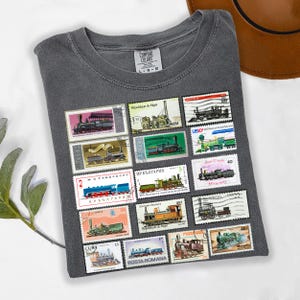 May include: A gray Comfort Colors t-shirt featuring a collection of vintage postage stamps with train illustrations. The stamps are in various colors and sizes, with text in multiple languages. The shirt is a size L-G-G.