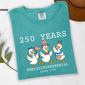 May include: Seafoam green t-shirt with a graphic of three cartoon ducks celebrating the 250th anniversary of the United States. The ducks wear patriotic accessories and hold an American flag. Text on the shirt reads "250 YEARS SEMIQUINCENTENNIAL SINCE 1776."