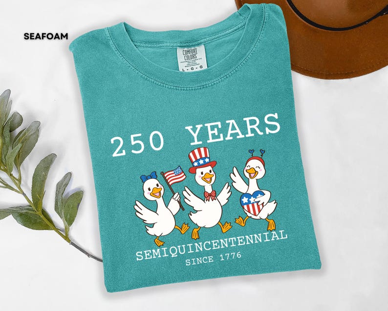 May include: Seafoam green t-shirt with a graphic of three cartoon ducks celebrating the 250th anniversary of the United States. The ducks wear patriotic accessories and hold an American flag. Text on the shirt reads "250 YEARS SEMIQUINCENTENNIAL SINCE 1776."