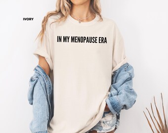 May include: Ivory-colored t-shirt with the text "IN MY MENOPAUSE ERA" in black. The shirt is paired with a light blue denim jacket. The shirt is a casual, comfortable style.