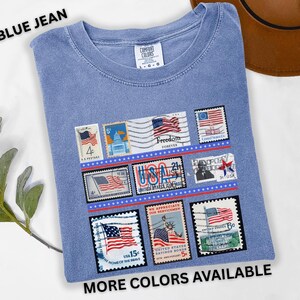 May include: Blue jean colored t-shirt with a graphic of vintage-style postage stamps featuring American flags and patriotic imagery. The shirt has the text "BLUE JEAN" and "MORE COLORS AVAILABLE".