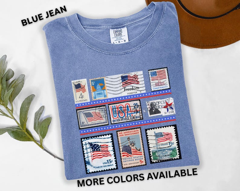May include: Blue jean colored t-shirt with a graphic of vintage-style postage stamps featuring American flags and patriotic imagery. The shirt has the text "BLUE JEAN" and "MORE COLORS AVAILABLE".