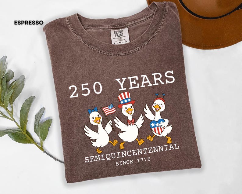 May include: Espresso-colored t-shirt featuring a graphic of three cartoon ducks celebrating with American flag imagery. The text reads "250 YEARS SEMIQUINCENTENNIAL SINCE 1776". The shirt is a Comfort Colors brand.