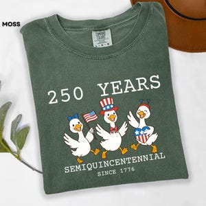 May include: Moss green t-shirt with a graphic celebrating 250 years. The design features three cartoon ducks wearing patriotic accessories and holding flags. Text includes "250 YEARS", "SEMIQUINCENTENNIAL", and "SINCE 1776".