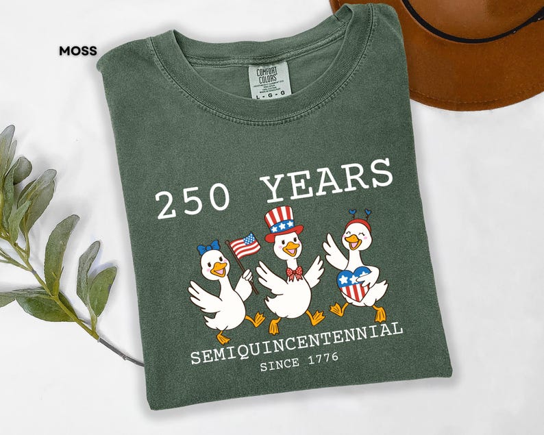 May include: Moss green t-shirt with a graphic celebrating 250 years. The design features three cartoon ducks wearing patriotic accessories and holding flags. Text includes "250 YEARS", "SEMIQUINCENTENNIAL", and "SINCE 1776".