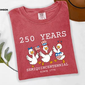 May include: Crimson red t-shirt with the text "250 YEARS SEMIQUINCENTENNIAL SINCE 1776." Features a graphic of three cartoon ducks wearing patriotic hats and holding flags. The shirt is a Comfort Colors brand.
