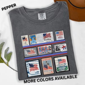 May include: Pepper-colored t-shirt featuring a collection of vintage-style postage stamps with American flag designs. The stamps are arranged in rows and include the text "Freedom Forever" and "USA". The shirt also has the text "MORE COLORS AVAILABLE".