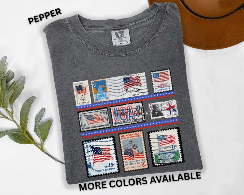 May include: Pepper-colored t-shirt featuring a collection of vintage-style postage stamps with American flag designs. The stamps are arranged in rows and include the text "Freedom Forever" and "USA". The shirt also has the text "MORE COLORS AVAILABLE".