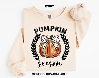May include: Ivory-colored sweatshirt with a pumpkin graphic. The design features a pumpkin with a polka-dot bow, surrounded by a wreath, and the words "PUMPKIN season". The text "MORE COLORS AVAILABLE" is also visible.