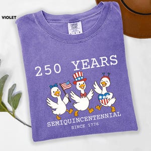 May include: Violet-colored t-shirt with the text "250 YEARS SEMIQUINCENTENNIAL SINCE 1776." Features a graphic of three cartoon ducks wearing patriotic hats and holding flags. The shirt is folded on a white surface.