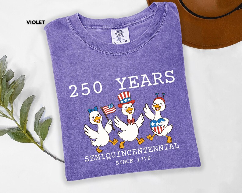May include: Violet-colored t-shirt with the text "250 YEARS SEMIQUINCENTENNIAL SINCE 1776." Features a graphic of three cartoon ducks wearing patriotic hats and holding flags. The shirt is folded on a white surface.
