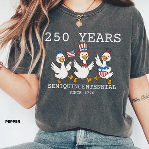 May include: Dark gray t-shirt with the text "250 YEARS SEMIQUINCENTENNIAL SINCE 1776." The shirt features a graphic of three cartoon ducks wearing patriotic accessories and holding American flags. The shirt is a casual style.