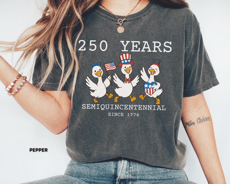 May include: Dark gray t-shirt with the text "250 YEARS SEMIQUINCENTENNIAL SINCE 1776." The shirt features a graphic of three cartoon ducks wearing patriotic accessories and holding American flags. The shirt is a casual style.
