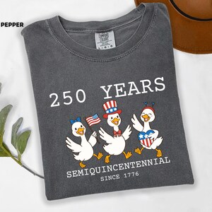 May include: Pepper-colored t-shirt with the text "250 YEARS SEMIQUINCENTENNIAL SINCE 1776." Features a graphic of three cartoon ducks in patriotic attire, holding flags and hearts. The shirt is a Comfort Colors brand.