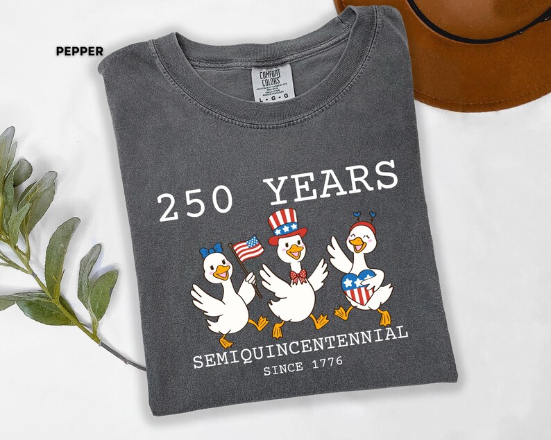 May include: Pepper-colored t-shirt with the text "250 YEARS SEMIQUINCENTENNIAL SINCE 1776." Features a graphic of three cartoon ducks in patriotic attire, holding flags and hearts. The shirt is a Comfort Colors brand.