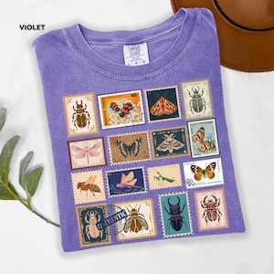 Może przedstawiać: Fioletowa koszulka Comfort Colors z kolekcją znaczk&oacute;w pocztowych w stylu vintage z ilustracjami owad&oacute;w. Znaczki przedstawiają r&oacute;żne owady, w tym chrząszcze, motyle i pszczoły. Na jednym ze znaczk&oacute;w znajduje się napis "AUTHENTIC".