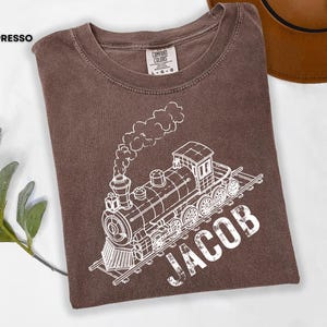 Puede incluir: Camiseta color espresso con una ilustración blanca de un tren antiguo y el nombre "JACOB" impreso debajo. La camiseta es de material suave y tiene un diseño clásico de cuello redondo. Un sombrero marrón es visible en el fondo.