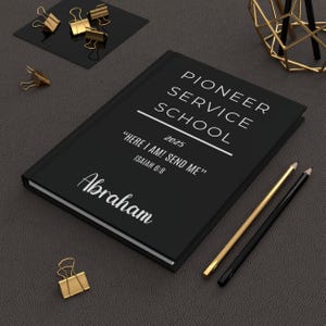Puede incluir: Un cuaderno negro con el texto "PIONEER SERVICE SCHOOL 2025" y la cita "HERE I AM! SEND ME" ISAIAH 6:8. El nombre "Abraham" está impreso en la portada. También se ven lápices dorados y negros y clips dorados.