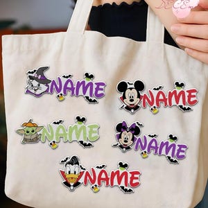 Peut inclure: Un sac fourre-tout en toile couleur crème orné de cinq écussons brodés portant le mot NAME en différentes couleurs. Les écussons représentent des personnages de dessins animés en costumes d'Halloween, dont un chat, Mickey, Grogu, Minnie et Donald.