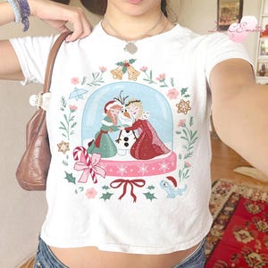 May include: White t-shirt featuring a snow globe design with the characters Anna, Elsa, and Olaf from the movie Frozen. The design includes floral and snowflake accents, a candy cane, and a small blue dragon.