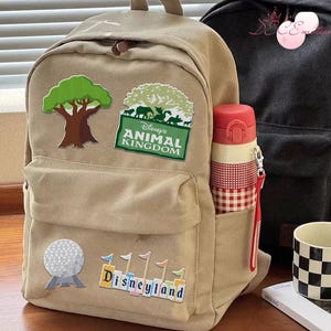 May include: Beige backpack featuring Disney's Animal Kingdom and Disneyland patches. A red and white water bottle is in the side pocket. A black backpack and a black and white chequered mug are also visible.