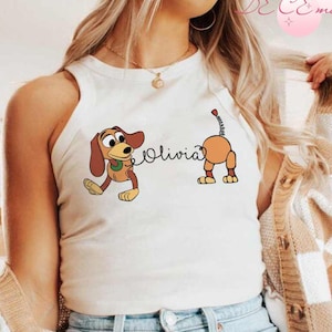 May include: A white tank top featuring a cartoon illustration of a dachshund dog with the name "Olivia" in cursive. The dog is brown and tan with a green collar. The tank top is sleeveless.