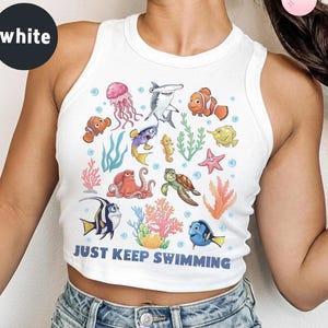 May include: White tank top with a colorful graphic of various sea creatures, including fish, a shark, and a jellyfish. The phrase "JUST KEEP SWIMMING" is printed in blue at the bottom of the design. The top is sleeveless and has a round neckline.