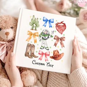 Peut inclure: Un livre photo à couverture rigide blanche avec un design coloré représentant un personnage de dessin animé, des bottes de cowboy, des nœuds, un cactus et un chapeau de cowboy. Le texte "Hardcover Photo Book" est en haut et "Custom Text" en bas.