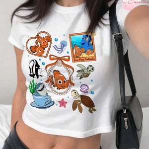 May include: White crop top with a colorful graphic print of characters from the movie Finding Nemo. The graphic includes Marlin, Dory, Nemo, and other sea creatures. The text on the graphic says "Just Keep Swimming".
